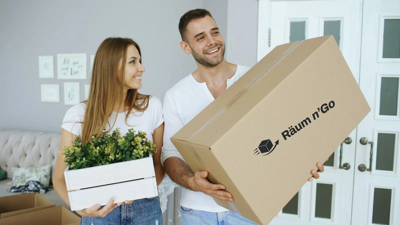Paar trägt einen Umzugskarton mit Räum n'Go Logo in die neue Wohnung